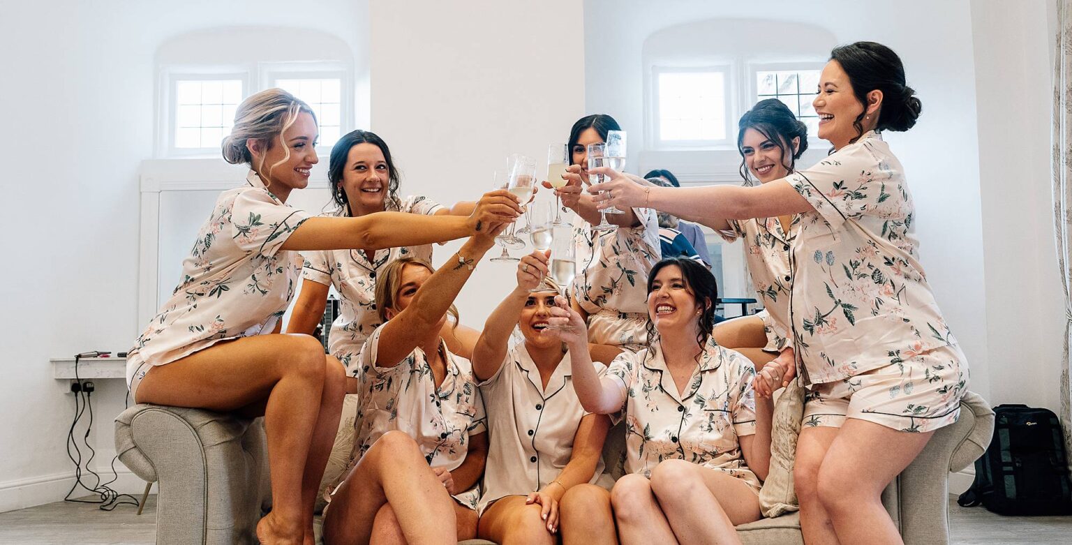 bridesmaids sit on sofa with bride and celebrate the wedding day with glasses of prosecco