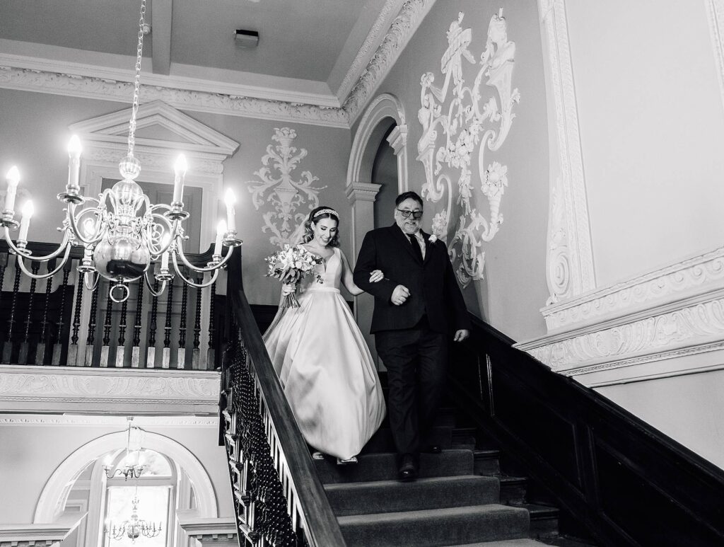 Black & white photo of bride walking down the staircase at Crowcombe Court with father of bride